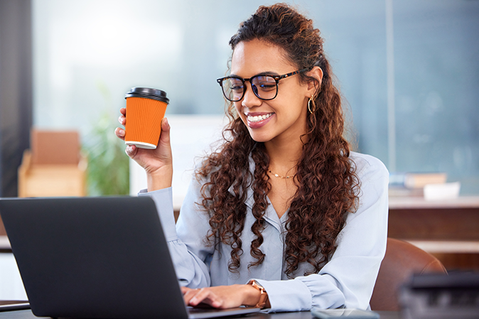 Young woman smiling while holding a coffee and working on laptop at her desk.